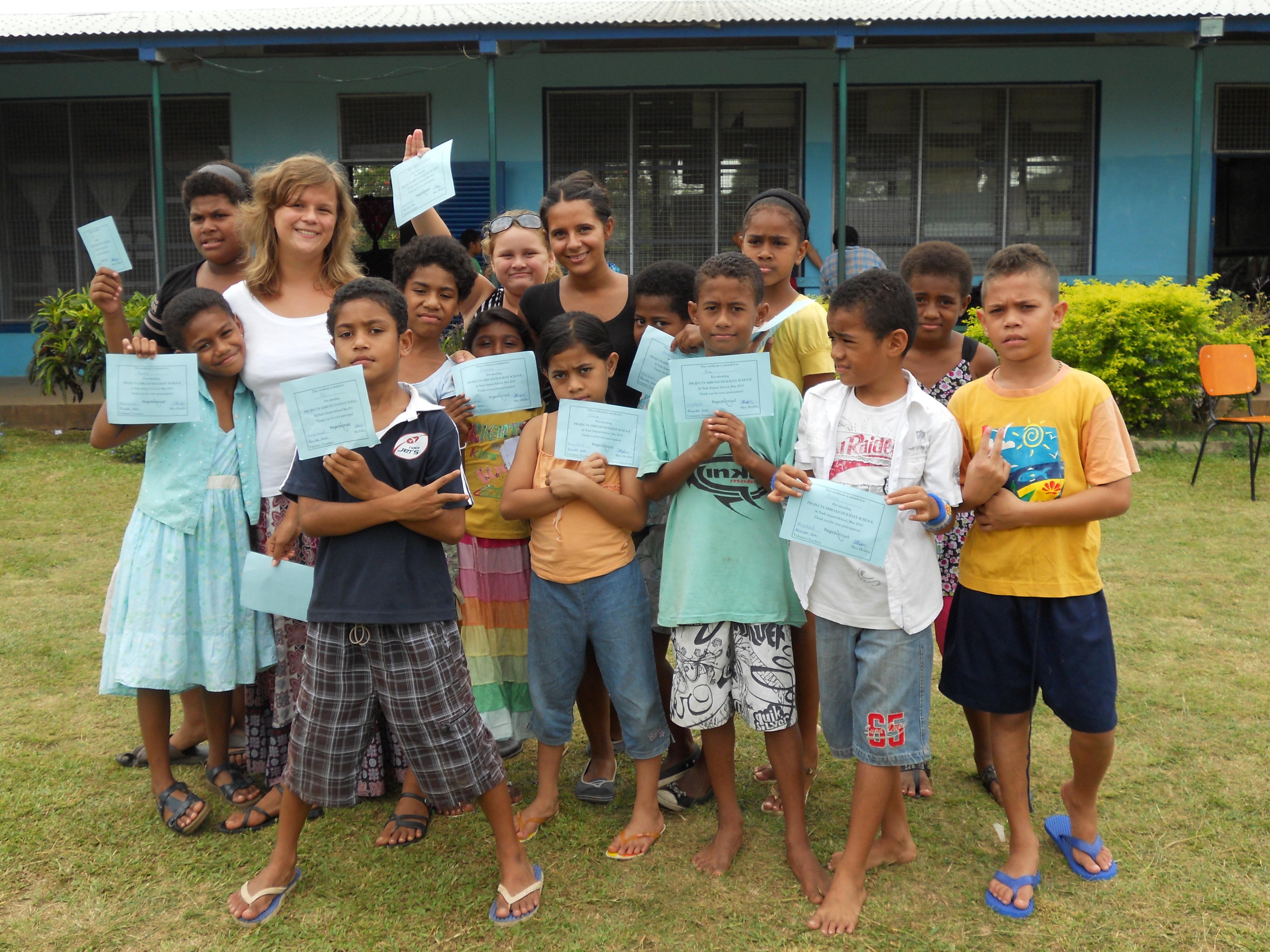 A volunteer teaching in Fiji poses for a photo with her students in Fiji A group photo of a Projects Abroad volunteer with her students outside her volunteer teaching placement in Fiji